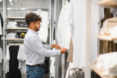 Genç Arap adam bir moda mağazasında geziniyor, şık erkek giyim seçeneklerinden şık bir beyaz gömlek seçiyor.