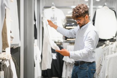 Şık bir adam giyim mağazasında geziniyor, beyaz gömleği dikkatle inceliyor ve şık bir gardırop için seçeneklerini değerlendiriyor.