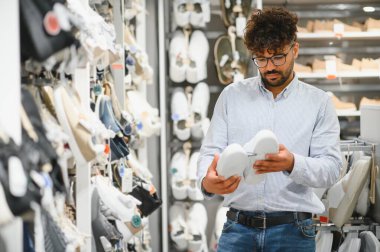 Zarif Arap adam modern bir ayakkabı mağazasında beyaz ayakkabıları dikkatlice seçiyor, alışveriş deneyiminin tadını çıkarıyor.