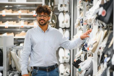 Genç Arap adam bir moda mağazasında ayakkabı alışverişi yapıyor, çeşitli stilleri inceliyor ve mükemmel uyumu arıyor.