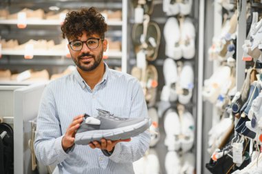 Genç bir Arap spor mağazasında koşu ayakkabısı seçiyor, vitrindeki şık seçeneklerin tadını çıkarıyor.