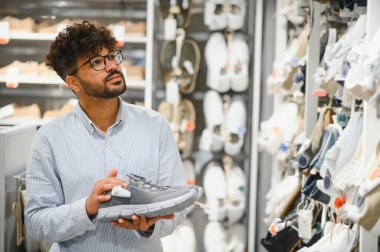 Sakallı Arap müşteri spor ayakkabı tutuyor, dik dik bakıyor, düşünceli bir şekilde ayakkabı seçerken seçeneklerle dolu geniş bir ayakkabı mağazasında