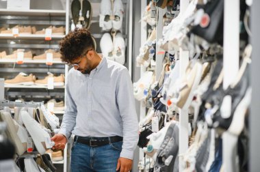 Genç Arap adam büyük bir moda mağazasında bir çift ayakkabı seçiyor, fiyat etiketlerine bakıyor ve farklı stilleri karşılaştırıyor.