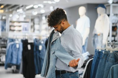 Genç, modaya uygun bir Arap adam modern bir giyim mağazasına bakarken şık bir kot ceket seçiyor ve rahat bir alışveriş deneyimi yaşıyor.