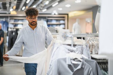Delikanlı Arap bir moda mağazasında çeşitli şık kıyafetleri karıştırıyor. Kişisel tarzını yansıtan giysileri dikkatlice seçiyor.
