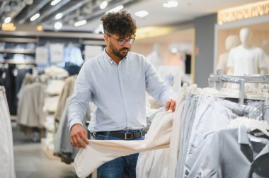 Zarif bir Arap adam giyim mağazasında şık pantolonlar seçiyor, alışveriş deneyiminin tadını çıkarıyor.