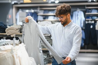 Sakallı Arap müşteri elinde gri gömlek, modern alışveriş merkezinde alışveriş kıyafetleri, hafta sonu indirimlerinin keyfini çıkarıyor.