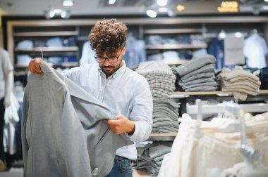 Gri ceketli genç bir Arap elbise mağazasında alışveriş yaparken gardırobu için seçenekleri değerlendiriyor.