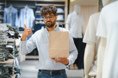Mutlu modaya uygun bir Arap adam, alışverişi ve büyük anlaşmalarıyla tatmin olmuş bir alışveriş çantasını elinde tutarken elini havaya kaldırıyor.