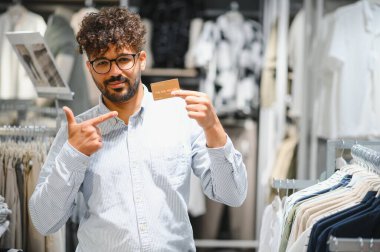 Kendine güvenen bir Arap, canlı bir perakende mağazasında şık kıyafetler alırken kredi kartını işaret ederek modern tüketici deneyiminin tadını çıkarıyor.