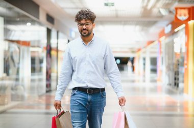 Arap adam parlak bir alışveriş merkezinden geçiyor, alışveriş torbaları taşıyor ve başarılı alışveriş deneyiminin tadını çıkarıyor.