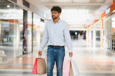 Arap adam alışveriş merkezinde elinde poşetlerle geziyor, başarılı alışveriş çılgınlığının tadını çıkarıyor.