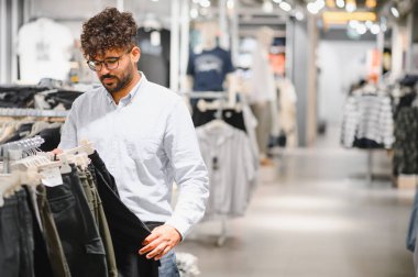 Genç Arap adam bir giyim mağazasında şık pantolonlar seçiyor, çeşitli seçenekleri incelerken zevkli alışveriş deneyiminin tadını çıkarıyor.