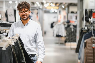 Gülümseyen Arap adam moda mağazasında alışveriş yapıyor, şık kıyafetler seçiyor ve indirimlerden faydalanıyor.