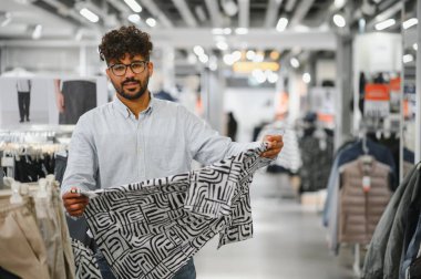 Genç Arap bir adam şık bir desenli gömleği incelerken bir giyim mağazasında geziniyor, sakin bir alışveriş deneyimi yaşıyor.