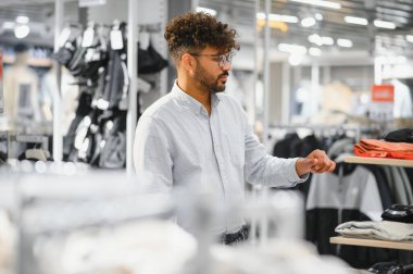 Arap adam modern bir alışveriş merkezinde elbise raflarını karıştırıyor, yeni moda eşyalar almanın zevkini çıkarıyor.