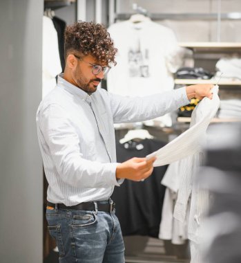Genç Arap bir adam şık bir gömleği incelerken bir giyim mağazasına göz atıyor, modern bir butikte sakin bir alışveriş deneyimi yaşıyor.