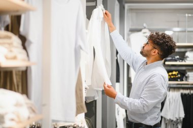 Arap bir adam giyim mağazasında dolaşırken beyaz bir gömleği kontrol ediyor, şık ve şık bir gardırop seçimi seçeneklerini değerlendiriyor.