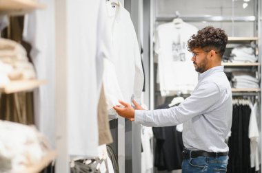 Modern bir alışveriş deneyimi yaşarken şık bir beyaz gömlek seçen şık bir Arap müşterisi moda mağazasında geziniyor.