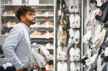 Tarz sahibi Arap adam büyük bir moda mağazasında ayakkabı seçiyor, çeşitli ayakkabılarla raflara bakıyor.