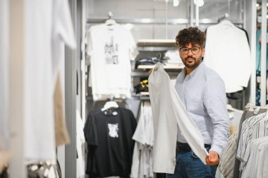Genç Arap bir adam modern bir giyim mağazasına bakarken şık bir beyaz gömleği inceliyor, sakin bir alışveriş deneyimi yaşıyor.