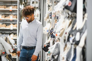 Genç, modaya uygun Arap bir adam bir ayakkabı mağazasında geziniyor, şık kostümünü tamamlamak için mükemmel ayakkabıları seçiyor.