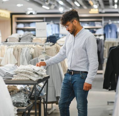 Bir giyim mağazasında kıyafetleri karıştıran şık bir adam moda kıyafetleri ve aksesuarları seçerken alışveriş deneyiminin tadını çıkarıyor.