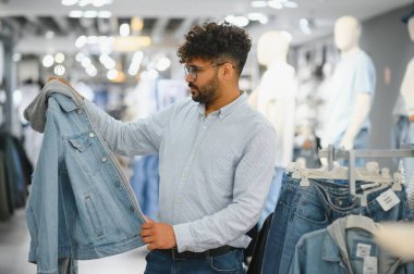 Sakallı Arap adam moda mağazasında şık kıyafetler seçerken elinde modaya uygun bir kot ceket tutuyor ve modern bir alışveriş deneyiminin tadını çıkarıyor.