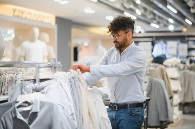Arap adam giyim mağazasındaki bir askıda kıyafetleri karıştırıyor, yeni moda eşyalar arıyor.