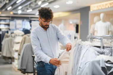 Genç Arap adam bir moda mağazasında kıyafet seçiyor, bir pantolon tutuyor ve kumaşın kalitesine bakıyor.