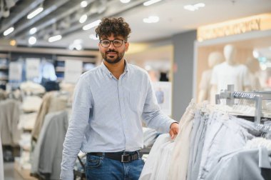 Şık bir adam giyim mağazasından gömlek seçiyor, canlı, modern bir alışveriş merkezinde zevkli bir alışveriş deneyimi yaşıyor.