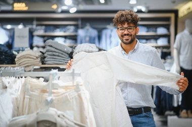 Mutlu genç Arap adam elbise alışverişi yapıyor, bir moda mağazasında beyaz gömlek tutuyor, alışverişinin tadını çıkarıyor.