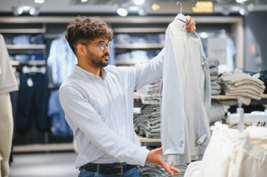 Sakallı Arap adam bir giyim mağazasında alışveriş yaparken açık mavi bir gömleği inceliyor, şık ve modern bir perakende deneyimi yaşıyor.