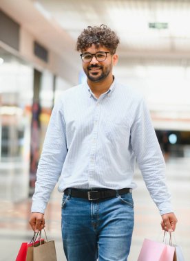 Alışveriş torbaları taşıyan genç bir Arap adam, alışverişlerinin keyfini çıkarırken ve gülümserken perakende heyecanıyla dolu canlı bir alışveriş merkezinde geziniyor.