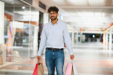 Arap adam modern bir alışveriş merkezinde yürüyor, alışveriş torbaları taşıyor ve mutlulukla dolu keyifli bir perakende terapi deneyimi yaşıyor.