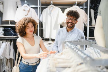 Gülümseyen bir çift, çeşitli seçeneklerle çevrili canlı bir moda mağazasında şık kıyafetler seçerken eğlenceli bir alışveriş deneyimi yaşıyor.