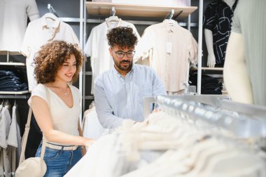 Gülümseyen çift canlı bir moda mağazasında elbise raflarını karıştırıyor, şık kıyafetler seçerken birlikte vakit geçiriyorlar.