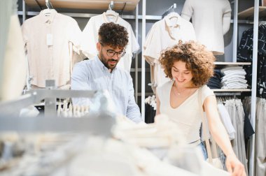 Neşeli bir moda mağazasında şık kıyafetleri karıştıran gülümseyen çift hafta sonları birlikte alışveriş yapmanın tadını çıkarıyorlar.