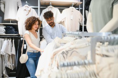 Genç Arap çift modern bir moda mağazasında şık giysi eşyasına bakıyorlar, alışveriş deneyimlerinin tadını çıkarıyorlar ve moda kıyafetlerini seçiyorlar.