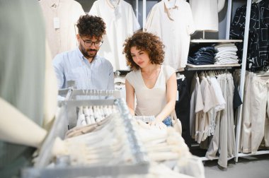 Genç Arap çift bir moda mağazasında çeşitli kıyafet seçeneklerini gözden geçiriyorlar, birlikte yeni kıyafetler seçmenin zevkini çıkarıyorlar.