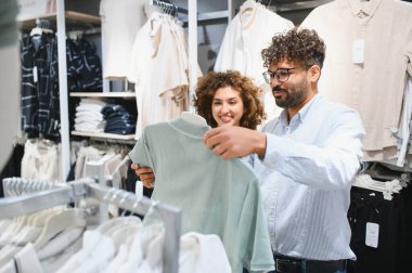 Mutlu Arap çift, canlı bir moda mağazasında dolaşırken ellerinde şık bir tişört tutarak alışveriş yapmaktan zevk alıyor.