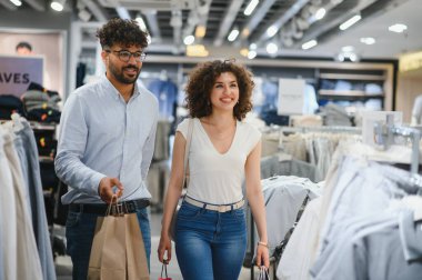 Gülümseyen Arap çift şık bir alışveriş çılgınlığının keyfini çıkarıyor, renkli çantalar taşıyor ve moda mağazalarında kıyafet seçeneklerini araştırıyor.