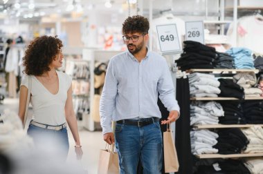 Arap çift bir giyim mağazasında geziyor, alışveriş torbaları taşıyor ve son zamanlardaki alışverişleri hakkında hararetli tartışmalar yapıyorlar.