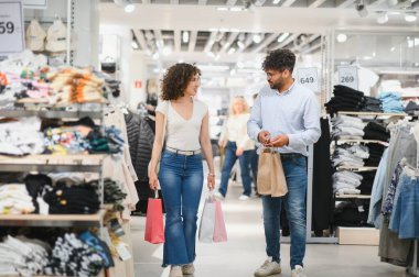 Mutlu çift moda mağazasında alışveriş yapıyor, renkli çantalar taşıyor ve alışverişleri tartışıyorlar.