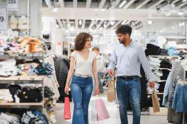 Mutlu çift birlikte vakit geçirmekten zevk alıyor, renkli alışveriş çantaları taşıyorlar ve canlı bir giyim mağazasında yürüyüp sohbet ediyorlar.