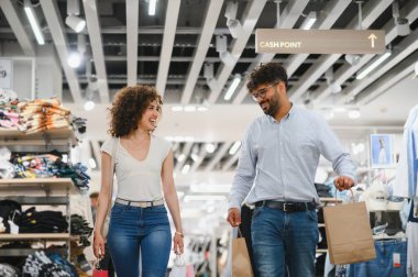 Gülümseyen bir çift şık bir kıyafet mağazasını keşfederken renkli kağıt poşetler taşıyarak neşeli bir alışveriş çılgınlığının tadını çıkarıyor.