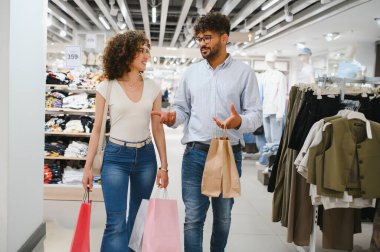Gülümseyen bir çift alışveriş torbaları taşıyarak moda mağazasında yürüyor, satışları tartışıyor ve alışveriş çılgınlığının tadını çıkarıyor.