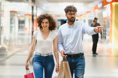 Mutlu bir çift alışveriş merkezinde yürürken, alışveriş torbaları taşırken adam şık bir mağazayı işaret ediyor.