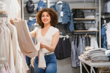 Neşeli bir moda mağazasında askılarda asılı şık kıyafetleri karıştıran gülümseyen genç bir kadın.