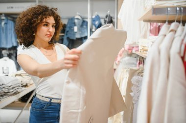 Müşteri bir moda mağazasında alışveriş yaparken beyaz ceketi inceliyor, yeni kıyafetler almanın zevkini çıkarıyor.